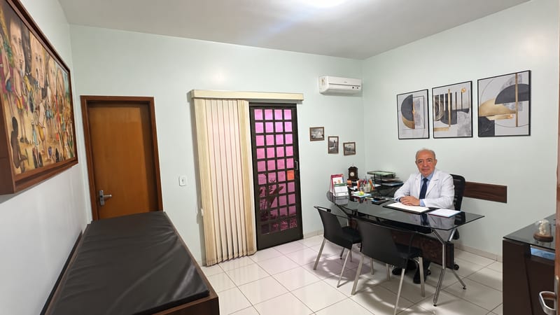 Consultório da Clínica Imune, com mesa de atendimento e maca, e o Dr. Paulo Prado à mesa, em Rio Verde